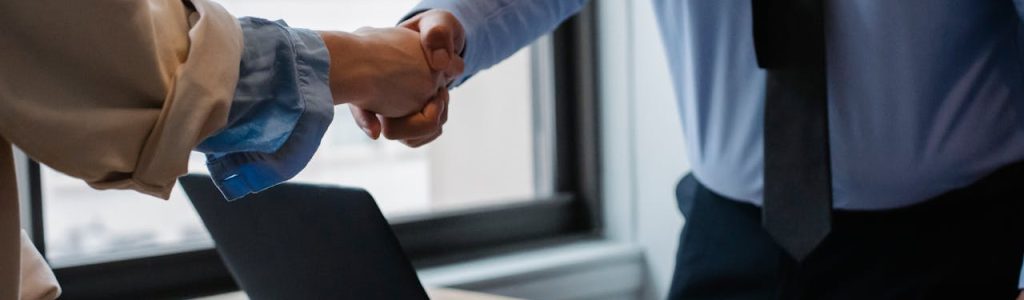 Crop unrecognizable coworkers in formal wear standing at table with laptop and documents while greeting each other before meeting